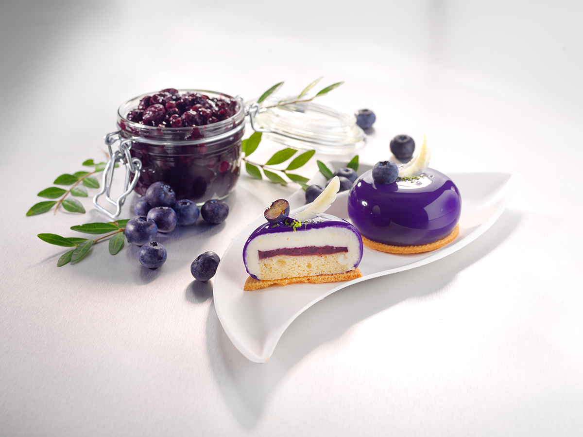 Photo d’un cheesecake, fourré à la myrtille, également composé d’un biscuit, d’une mousse au fromage blanc citron et d’un glaçage miroir violet.