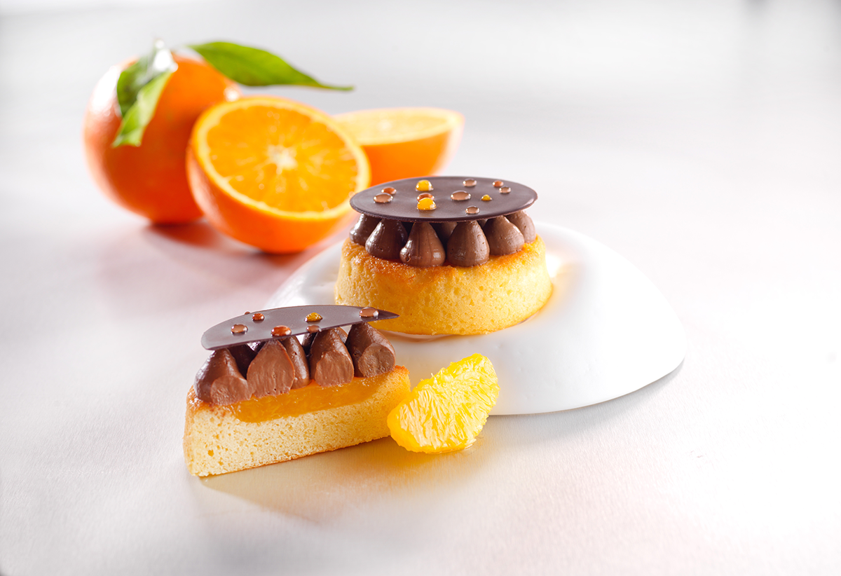 Photo d’un petit biscuit madeleine moelleux, fourré à l’orange, revêtu d’un crémeux chocolat noir poché à la douille et d’un disque au chocolat noir décoré de points de glaçage miroir coloré.