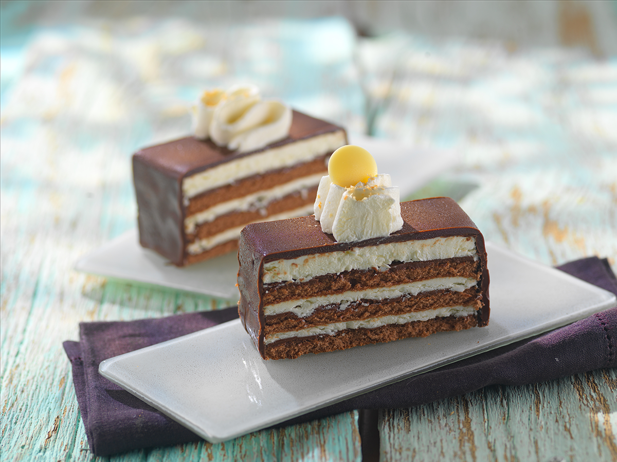 Photo d’une part d’un entremet alternant des couche de garniture cacao chocolat noisette et de fourrage vanille avec une finition glaçage cacao.