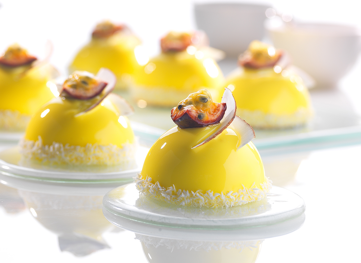 Photo d’un entremets individuel en forme de dôme avec un glaçage miroir jaune, composé d’une mousse à la pomme et d’une compotée à la passion.