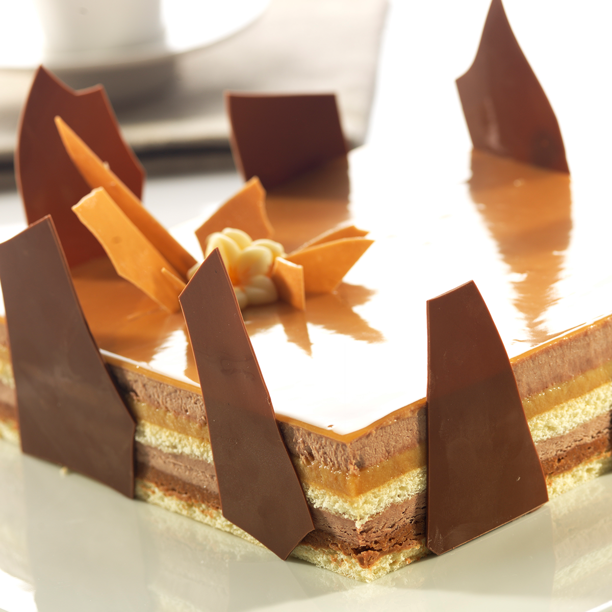 Photo d’un entremets carré avec une alternance de couches de mousse au  chocolat au lait, faite avec Bavaria chocolat lait, de la marque Dejaut, et au  caramel avec une finition faite de grandes brisures et éclats de chocolat au lait  et de caramel, et de Royal miroir caramel de la marque Marguerite